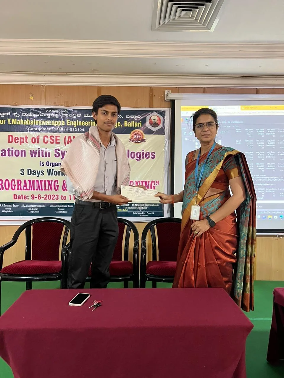 A student receiving a certificate on stage, with model training output displayed on the screen behind
