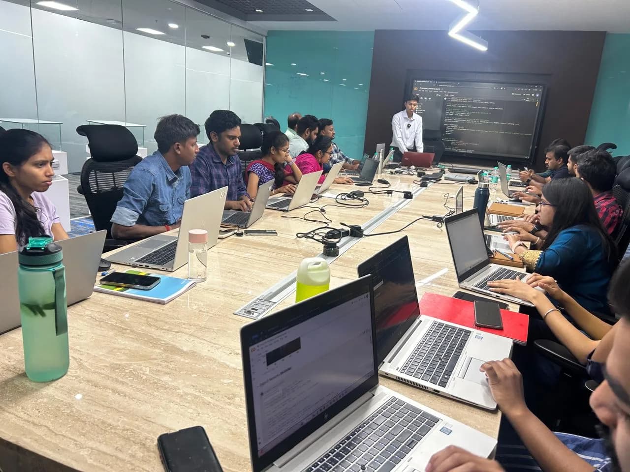 Instructor presenting code on a large screen to a boardroom of participants with laptops